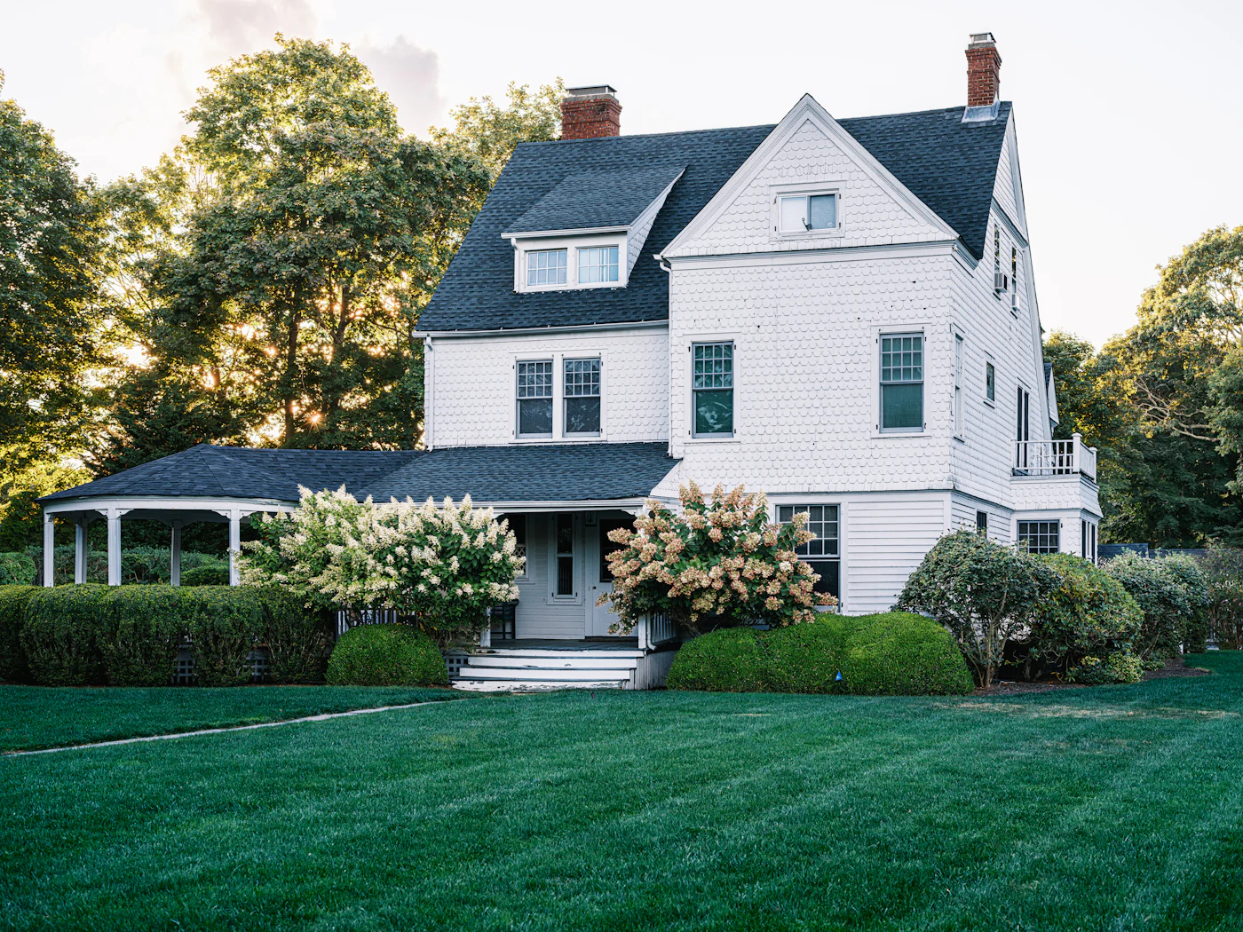 Full exterior of a shingle-style residence with a wraparound porch, mature trees and lawn